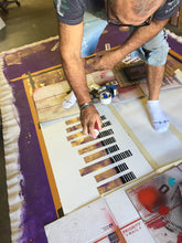 Maco is kneeling on the floor while spray painting black piano keys onto a white canvas. The workspace is covered in paint and art supplies, with a prominent purple paint stroke on the left.