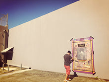 Marco stands next to a large Prince painting and gold memorial-style wall display mounted on a beige wall under a blue sky.