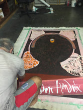 Marco kneels beside a large framed artwork on a workshop floor, applying or smoothing a pink strip along the bottom edge of the piece. The artwork features a dark central oval with pale pink side panels and white script text near the bottom.