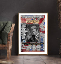 A framed David Bowie print leaning against a dark wall on a wooden floor, next to a green chair. The print features a black-and-white portrait of Bowie with red, white, and blue collage-style text and graphics around it, enclosed in a thin gold frame.