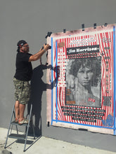 Marco stands on a metal step ladder against a gray wall, holding a camera  in front of a large poster for details shots. The poster features a black-and-white portrait of Jim Morrison with red, white, and blue striped borders and text elements.