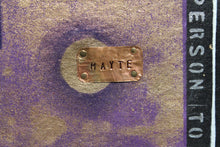 A close-up of a textured purple and gold fabric surface with a rectangular copper-toned metal nameplate stamped with the name 'MAYTE' Prince  wife