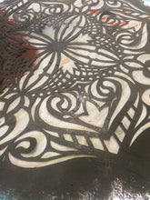 A close-up of a dark gray metal sheet featuring an intricate laser-cut filigree pattern over a white surface.