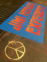 A close-up of a printed textile or mat featuring the words JIMI HENDRYX EXPERIMENT' in large red-orange lettering on a blue rectangular panel, with a gold peace-symbol-like emblem below.