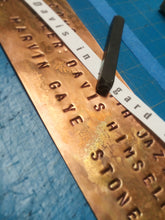 A close-up of a copper metal nameplate with raised letters spelling names, resting on a blue cutting mat with a black stamp.