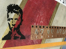 A close-up of a textured collage print featuring a black-and-gold stencil portrait of David Bowie, a diagonal red stripe with gold foil-like accents, a torn brown film strip with Alien  letters. 