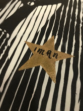 A close-up of a black and white vertically striped fabric featuring a central gold glitter star with the text IMAN written in black script.