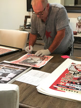Marco wearing a gray t-shirt with a red star and white gloves is signing  a print  laid out on a wooden table. The table is surrounded by other posters and papers in a home or studio setting.