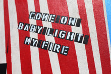 A close-up of a red and white striped textile with the phrase 'COME ON BABY LIGHT MY FIRE' printed in black block letters with white outlines.